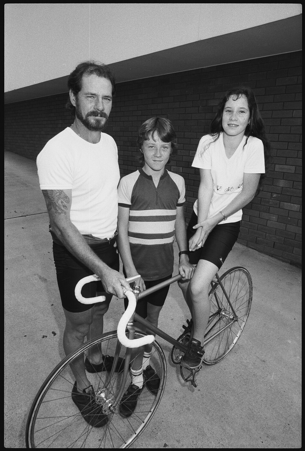 Unidentified man, boy, and girl with racing bicycle in car park outside the Queensland Times office, West Ipswich, January 1986