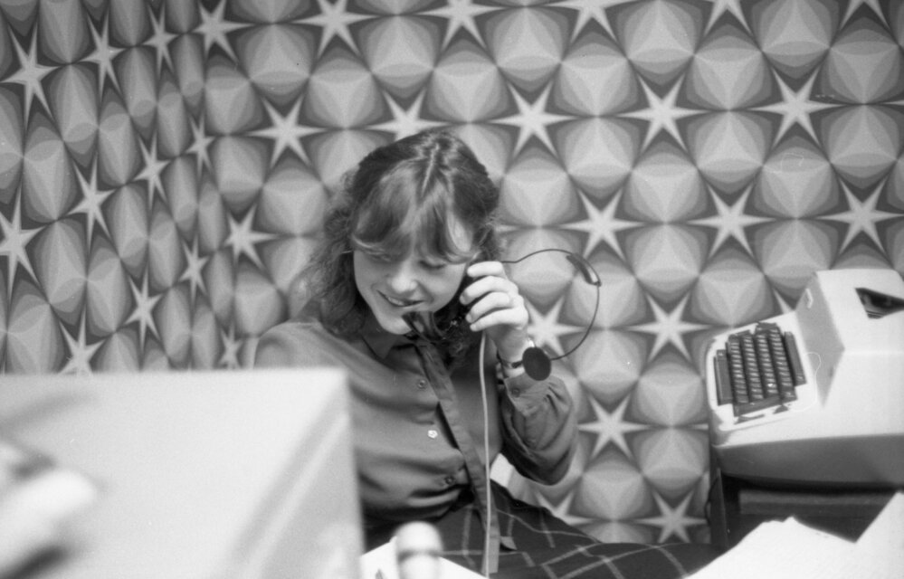 Unidentified young woman speaking into a phone headset while working at a desk, Ipswich, August 1980