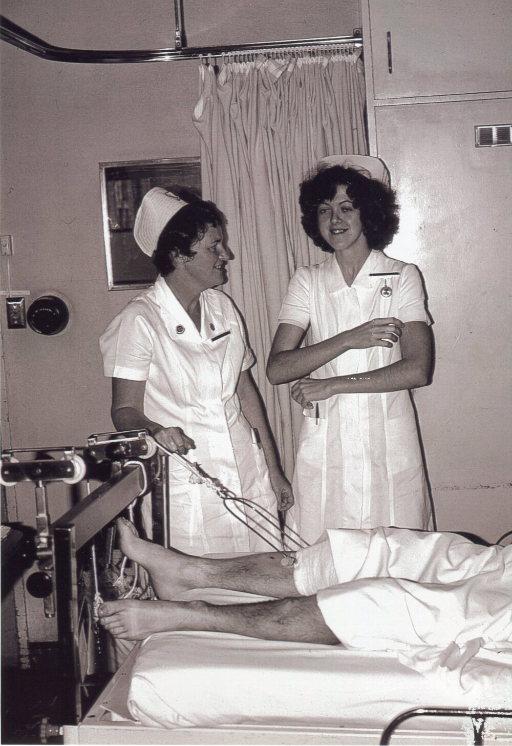 Orthopaedic patient in traction, with nurses Jan Wilton (nee White) and Jenny Moore, Ipswich Hospital, 1970s