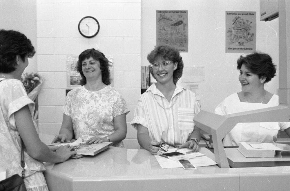 Unidentified staff members at Redbank Plaza Library helping a patron check out a book, Redbank, September 1985