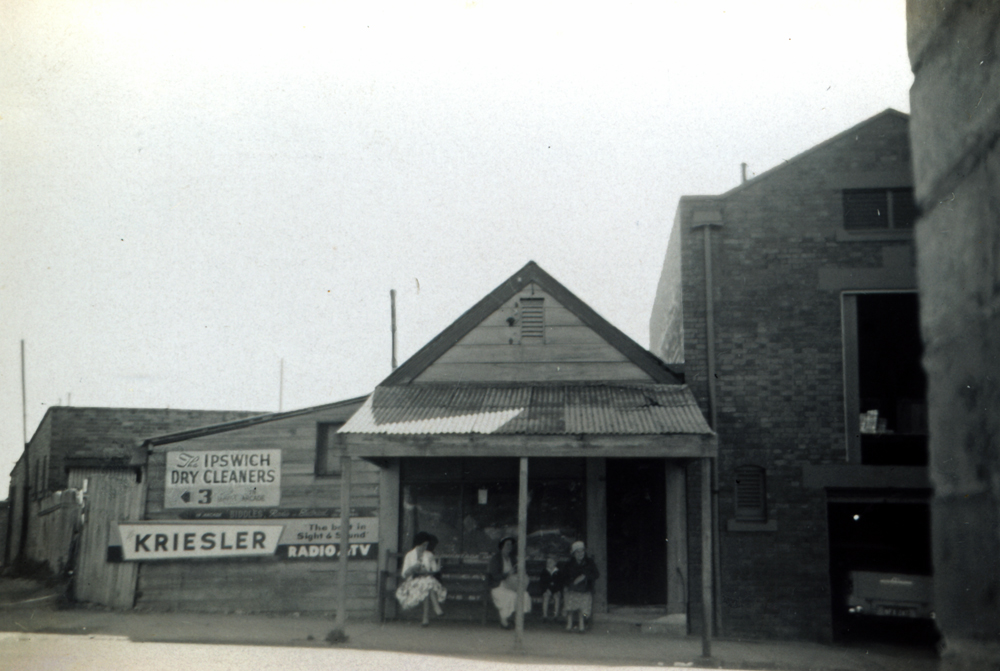Three women and child sitting on seat in front of W H Bemi, Printer, building, Ellenborough Street, Ipswich, 1952
