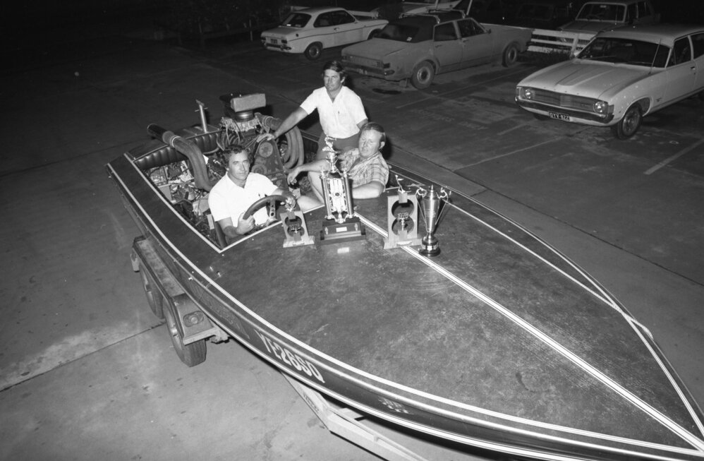Three unidentified men with trophies in a speedboat on a trailer in a carpark with cars, Ipswich February 1978