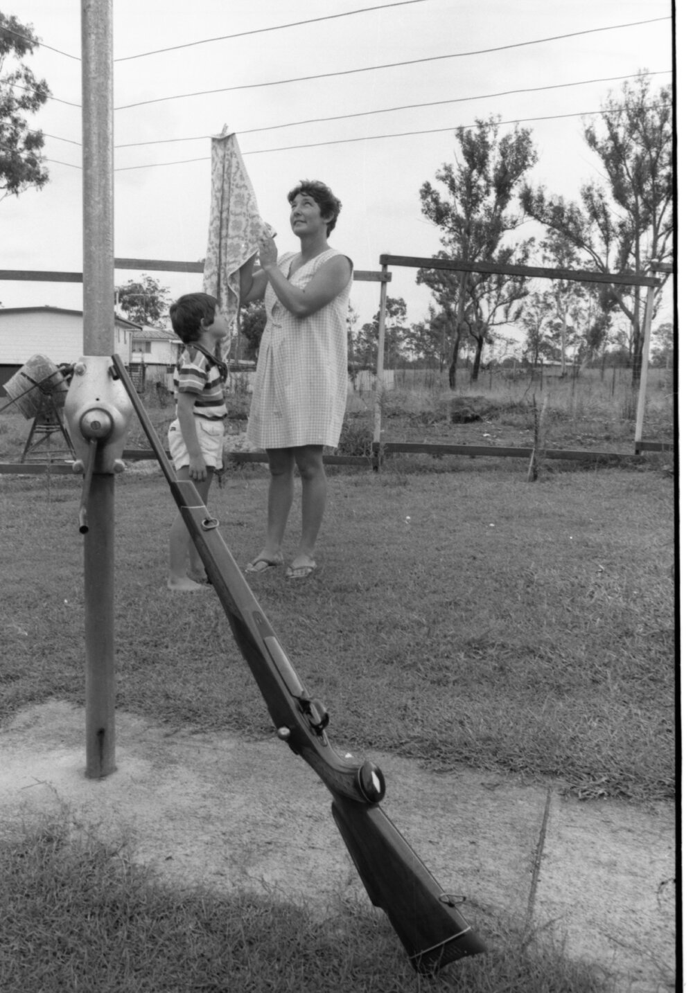 Unidentified woman and child in backyard hanging up laundry on a Hills Hoist washing line with rifle, Ipswich, February 1978