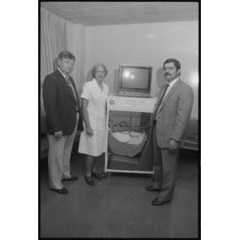 Dr Vlas Efstathis, a Nursing Sister and an unidentified man presenting a Ipswich Rotary Club project of a baby capsule, Ipswich, August 1985
