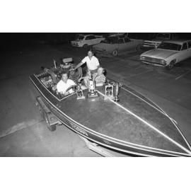 Three unidentified men with trophies in a speedboat on a trailer in a carpark with cars, Ipswich February 1978